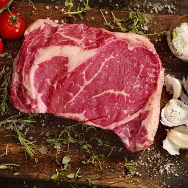 28-day dry aged Welsh rib eye steak on wooden board with herbs, garlic, salt, and tomatoes