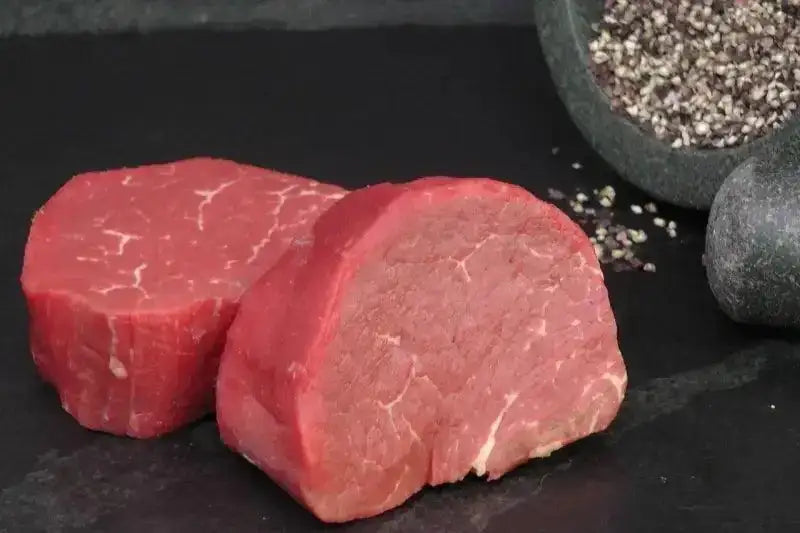 Raw premium fillet steaks on dark slate with cracked black pepper in a mortar