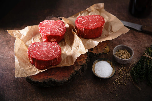 Three raw beef steaks on butcher paper with herbs, salt, and peppercorns on a rustic board