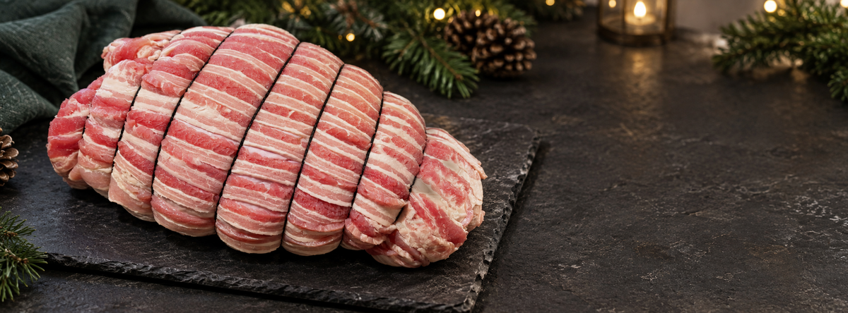A raw, rolled turkey wrapped in bacon-wrapped meat sits on a dark slate platter on a dark tabletop, with pine branches, a candle, and warm lights blurred in the background.