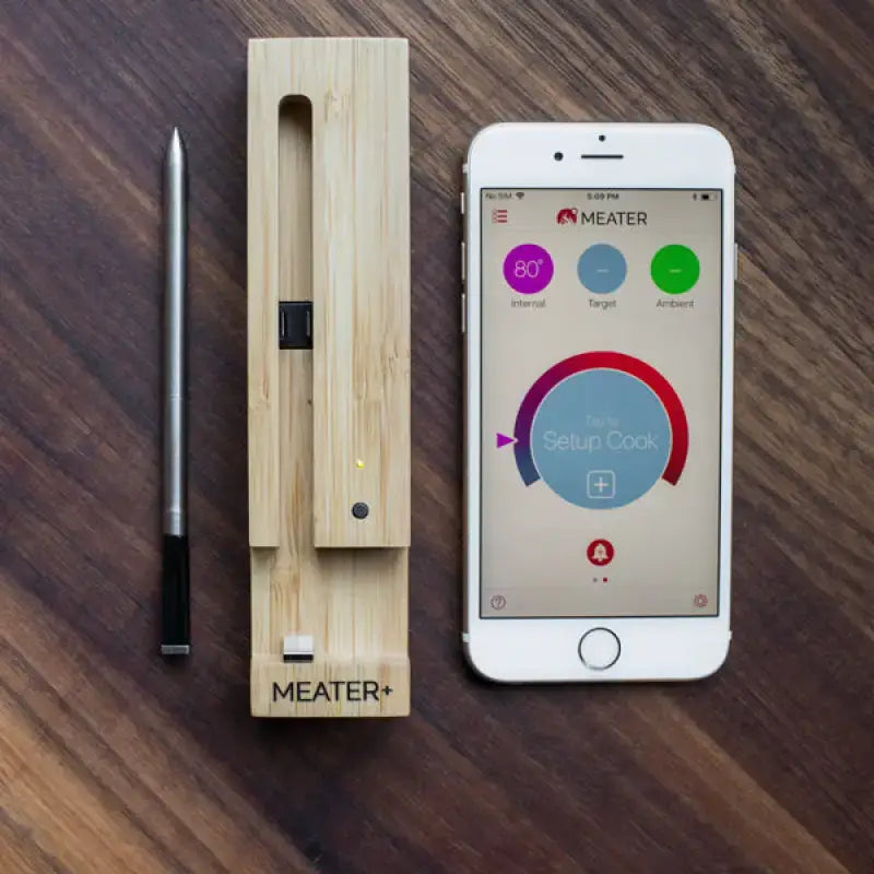 MEATER Plus Smart Thermometer with bamboo base and smartphone app displaying cooking settings on wooden surface.