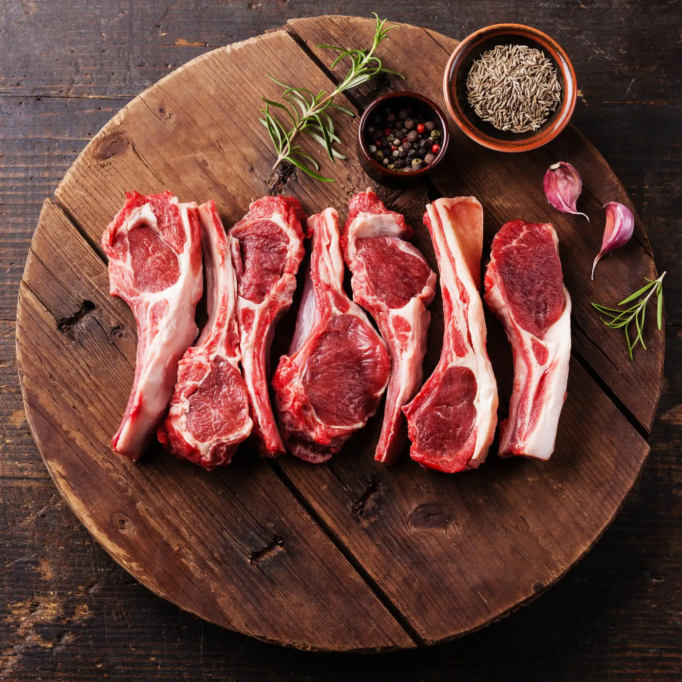 Raw lamb chops arranged in a row with herbs and spices on a wooden board.